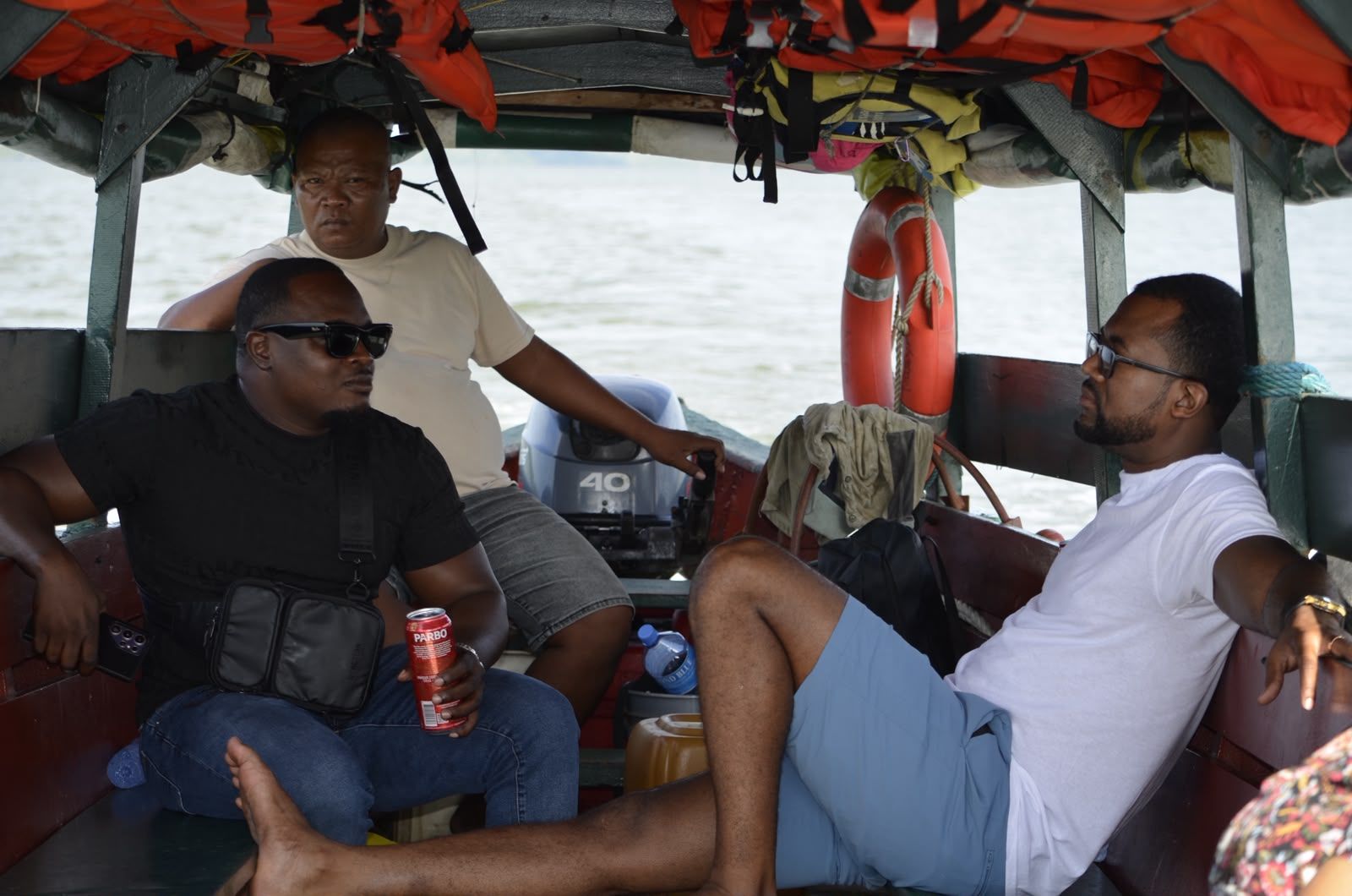 Drie mannen in een overdekte rivierboot: Andy leunt rechts ontspannen achterover, zijn vriend zit links met een blikje Parbo, de schipper zit achterin bij de buitenboordmotor. Zwemvesten hangen in het dak, een reddingsboei aan de zijkant.