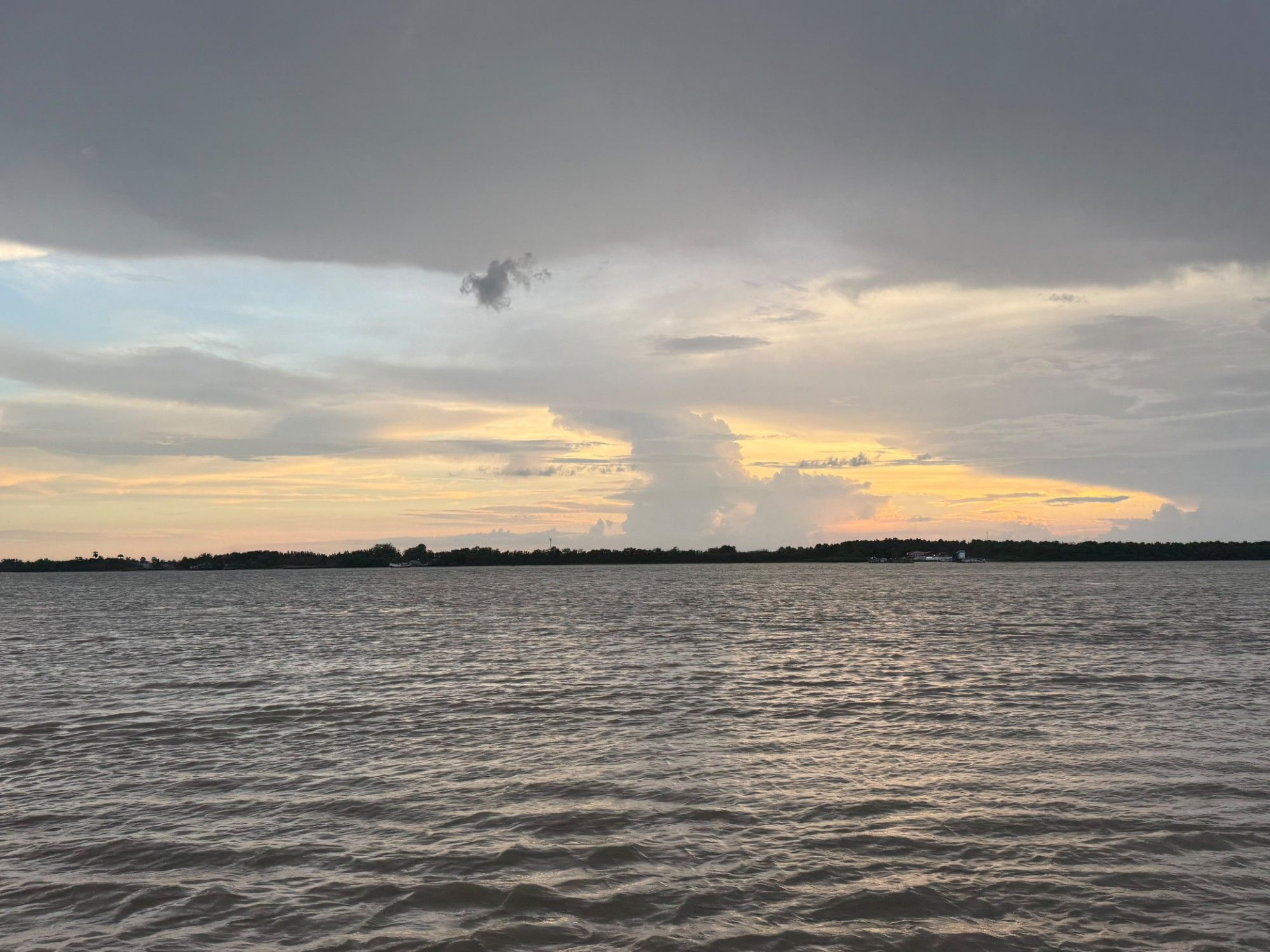 Zonsondergang boven de brede bruine rivier: een lage bosrand met een steiger aan de overkant, een stapelwolk in geel licht en een donkere wolkenlaag erboven.